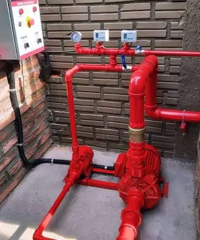 Casa de bombas de incêndio com duas bombas vermelhas, tubulação de recalque, manômetro, dois pressostatos instalados na tubulação e quadro de comando na parede.
